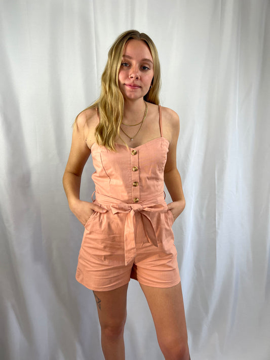 dusty pink linen blend romper with adjustable straps front button detail pockets and tie waist composed of cotton and spandex