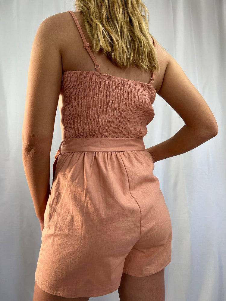 dusty pink linen blend romper with adjustable straps front button detail pockets and tie waist composed of cotton and spandex